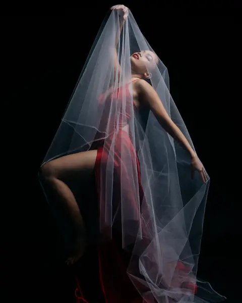 A studio photograph of a model in red beneath a translucent veil, against a black background