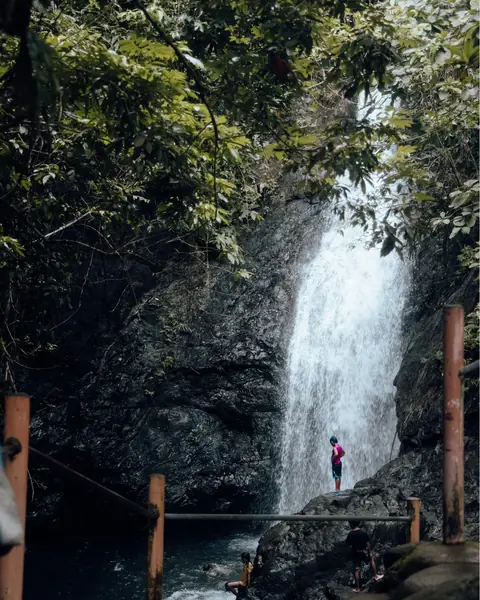 A white waterfall plunges down a dark rock face into a forest pool, with visitors showing its scale
